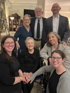Seven adults pose together in a living room; an elderly woman sits in a wheelchair at the center, surrounded by smiling relatives.
