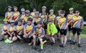 Group of cyclists wearing yellow and blue jerseys with helmets posing together outdoors on a gravel path.