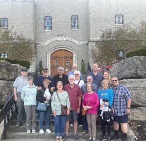 Group of adults posing on stone steps in front of a arched wooden door, ivy-covered walls behind them.