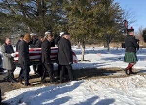 Outdoor funeral procession in a snowy park as six pallbearers carry a flag-draped casket; mourners follow behind.