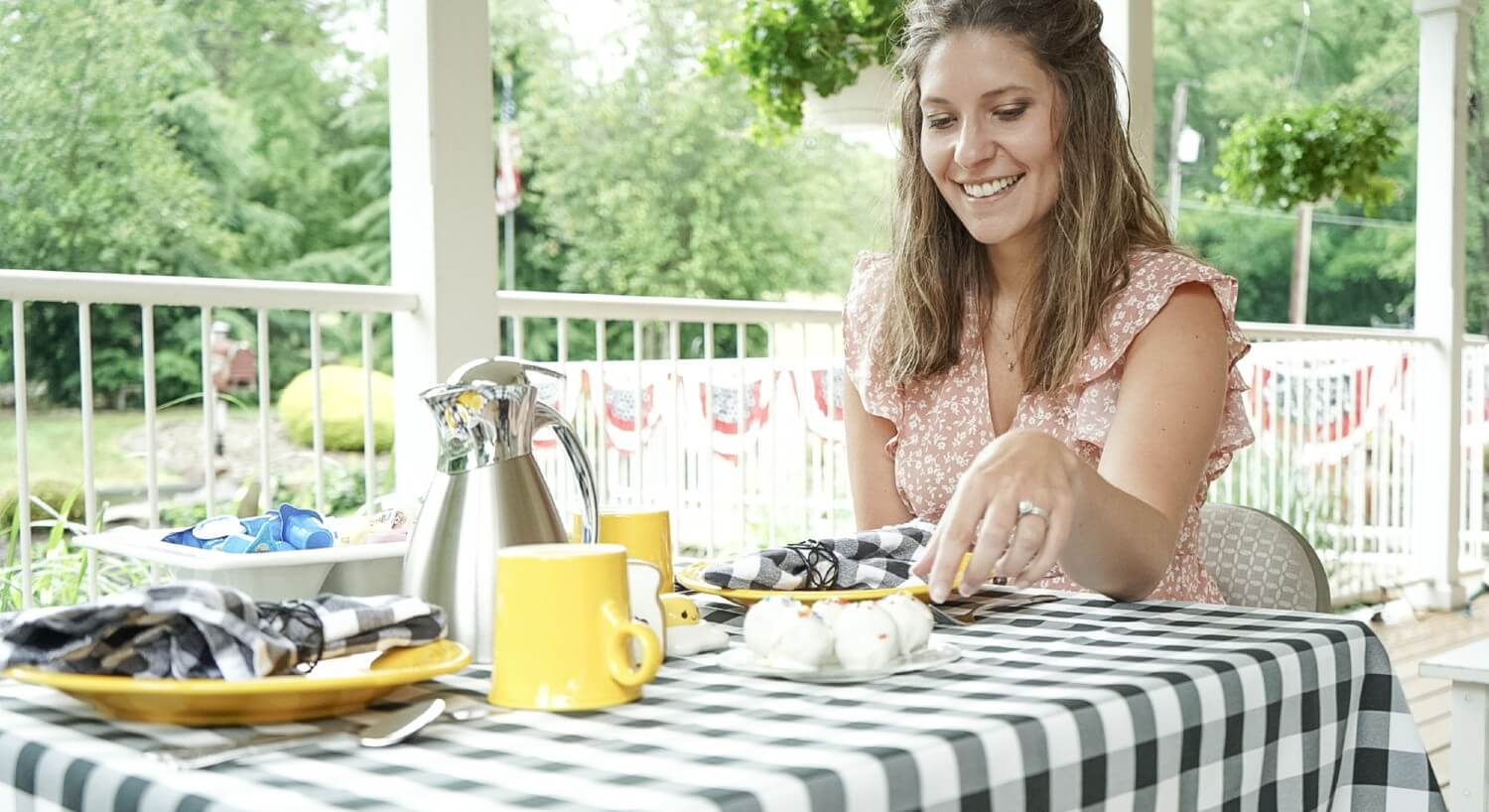 A smiling woman sitting at a table for two on an outside deck with trees in the background