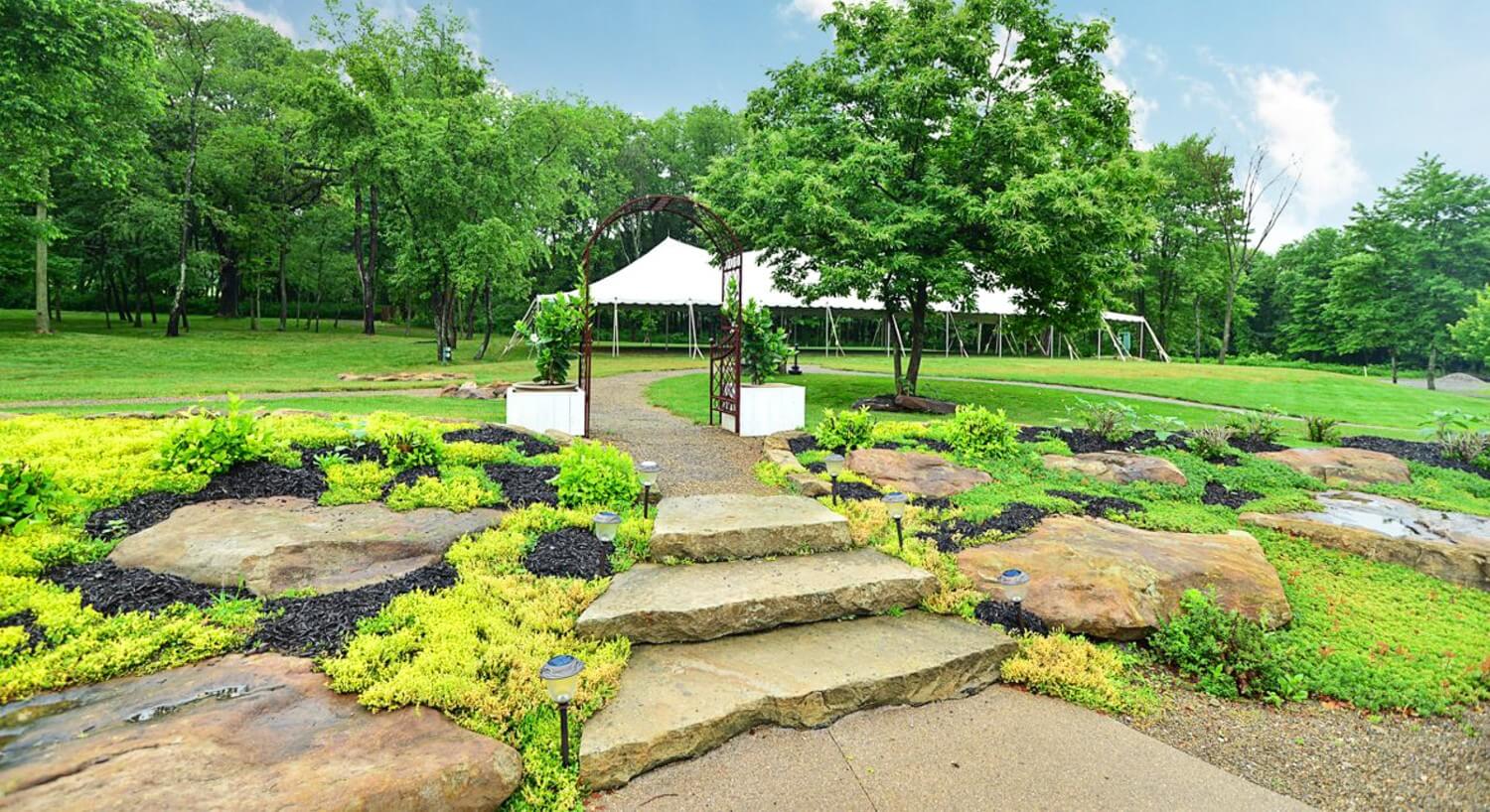 Decorative stone walkway leading up to an arch with an outdoor event tent and trees in the background