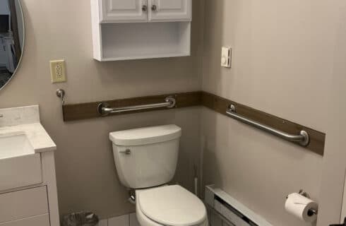 A bathroom with toilet, sink, mirror above the sink, and white cupboard above the toilet, along with grab bars.