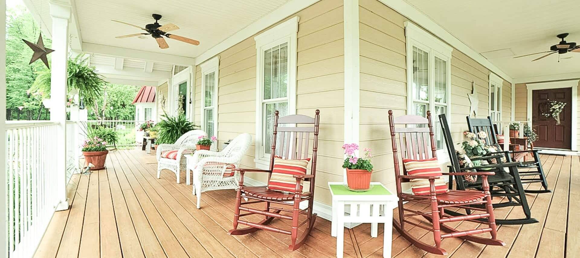 A large wrap around porch with white railings, four rocking chairs and two wicker chairs with a side table